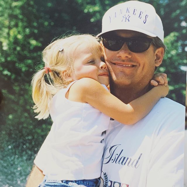 Gracie Dzienny's childhood photo in a white baby top hugging her father in a white t-shirt and a cap.